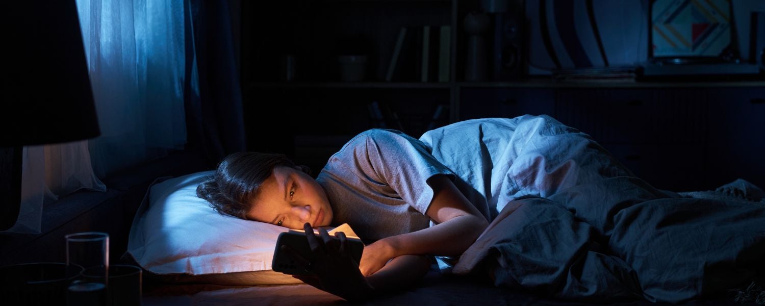 Personne allongée dans son lit la nuit, regardant un smartphone dont la lumière éclaire son visage dans une chambre sombre.