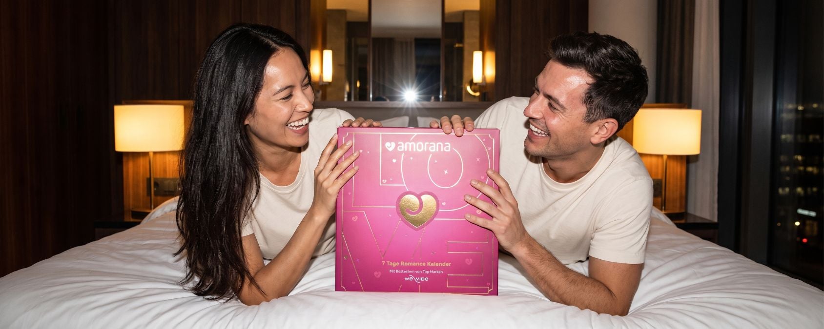Paar entdeckt gemeinsam den Amorana Romance Kalender 2026 auf dem Bett