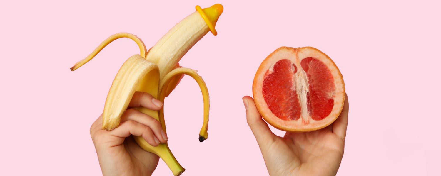Eine Hand hält eine Banane mit einem Kondom, eine andere Hand hält eine halbierte Grapefruit vor rosafarbenem Hintergrund.Show more lines