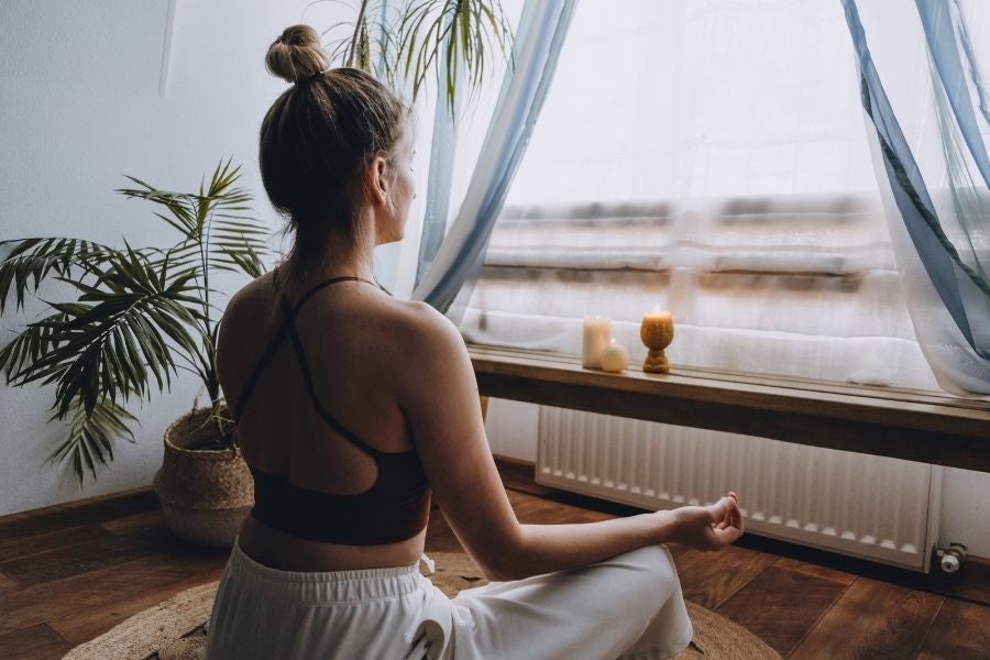 Achtsamkeit und innere Ruhe durch Meditation Person sitzt in einem hellen Raum auf einer Matte in Meditationshaltung vor einem Fenster. Pflanzen und Kerzen schaffen eine ruhige, achtsame Atmosphäre.