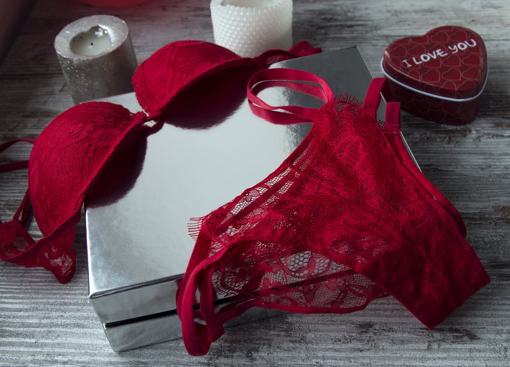Sinnliche Überraschung für einen besonderen Abend zu zweit Rote Spitzen-Dessous auf einer Geschenkbox neben Kerzen und einer herzförmigen Dose auf einem Holztisch.